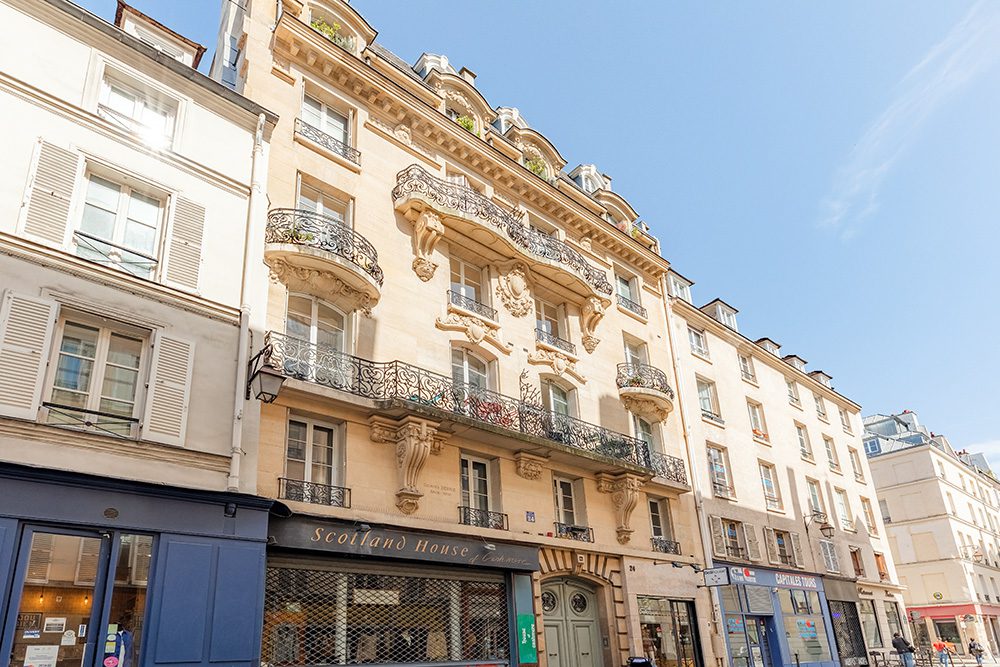 Parisian building with ornate wrought-iron balconies, decorative stonework, and storefronts along a sunny street.