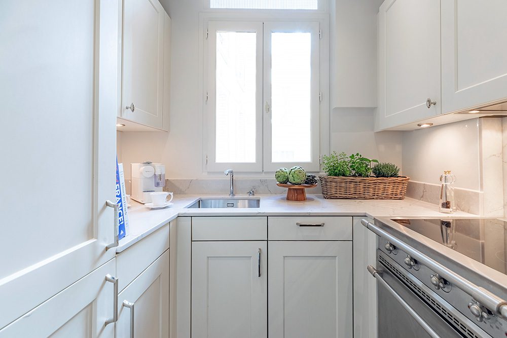 Roi de Sicile Paris fractional ownership Bright white compact kitchen with marble counter, window, sink, and plants in baskets.