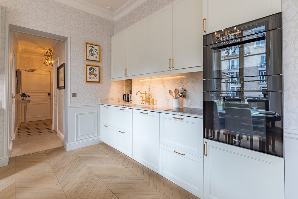 Tour du Pin Paris fractional ownership Bright white kitchen with gold hardware, marble backsplash, and built-in oven; hallway with wallpaper seen.